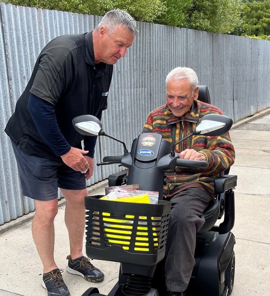 Mobility Centre staff, Gary, is dressed in his uniform. He leans forward to show Sir Robert Gillies how to drive his new scooter. Sir robert has short grey hair and wears a striped jumper.