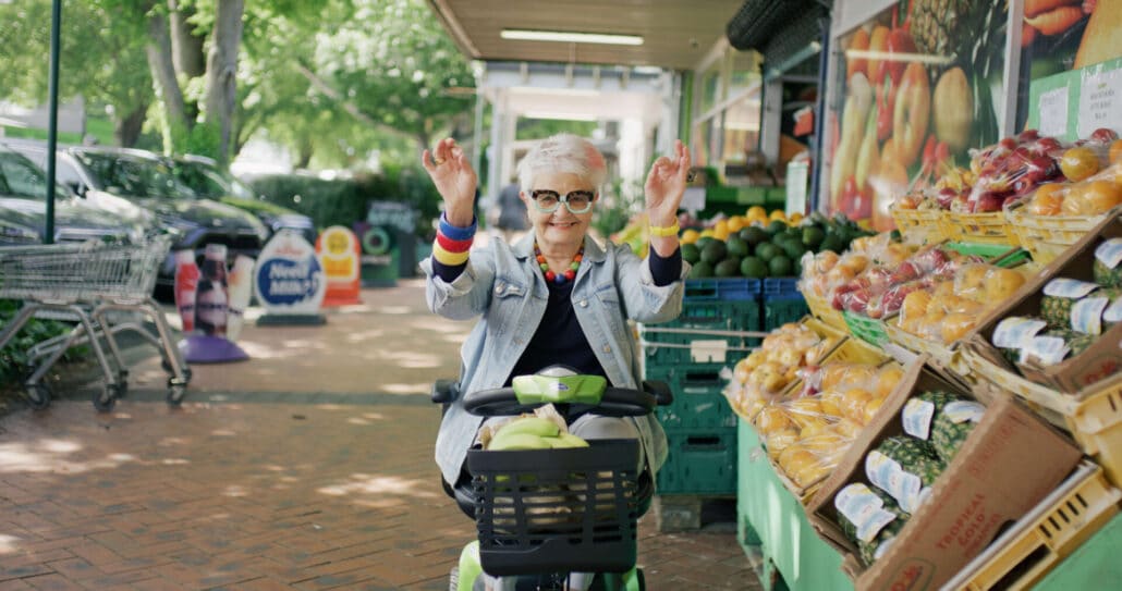 A woman is grinning as she rides her mobility scooter down a market street, past a green grocer.